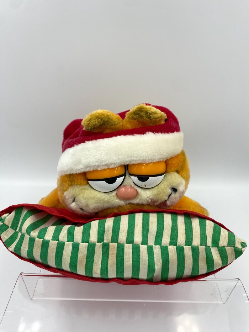 Może przedstawiać: Pluszowa zabawka Garfield w czerwono-białej czapce Świętego Mikołaja, spoczywająca na zielono-białej poduszce w paski z czerwoną obw&oacute;dką. Pomarańczowy kot ma r&oacute;żowy nos i senne oczy.
