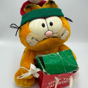 May include: A plush Garfield toy wearing a red and green knit hat with a white pom-pom. The orange cat is holding a red gift bag with the text "SHOP TIL YOU DROP". The bag has a green top and a white bow.