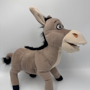 Könnte beinhalten: Ein Esel-Stofftier mit hellbraunem Fell, einer cremefarbenen Schnauze und schwarzen Hufen. Das Plüschtier hat eine schwarze Mähne, eine schwarze Schwanzquaste und große, ausdrucksstarke Augen. Es steht in einer Seitenansicht mit leicht geöffnetem Maul.