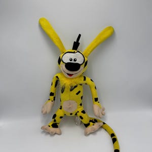 Peut inclure: Une peluche Marsupilami, une créature jaune avec des taches noires, de longues oreilles et une longue queue. Le jouet a un nez noir, des yeux et une petite touffe de cheveux noirs. Le jouet est sur une surface blanche.