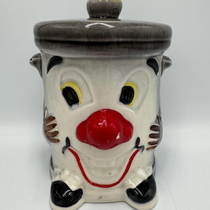 Può includere: Un barattolo per biscotti vintage in ceramica a forma di faccia da clown. Il barattolo ha una base bianca con un naso rosso, occhi neri e un sorriso rosso. Il coperchio e la maniglia sono grigio scuro. Ha piccole mani marroni e piedi neri.