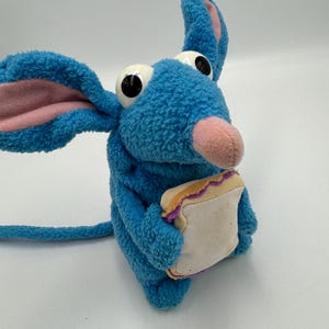 May include: A blue plush mouse toy with large eyes, a pink nose and ears. The mouse is holding a small sandwich with a white centre and a purple and yellow crust. The mouse has a long blue tail.