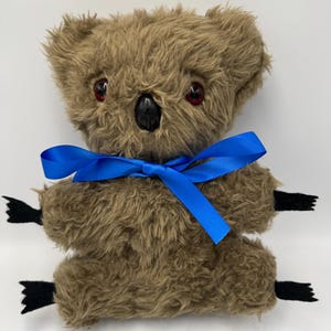 Può includere: Un peluche a forma di koala marrone con occhi rossi e naso nero. Ha un nastro blu e piedi in feltro nero. Il giocattolo è progettato per stare seduto. Il koala è realizzato in materiale morbido e soffice.