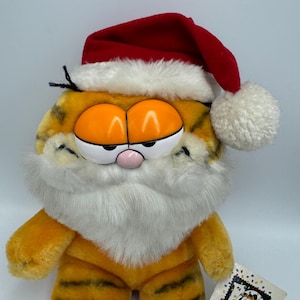 Può includere: Un peluche di Garfield con un cappello da Babbo Natale e una barba bianca. Il gatto arancione ha strisce nere e un naso rosa. Un'etichetta con l'immagine di Garfield e le parole "the party" è allegata.