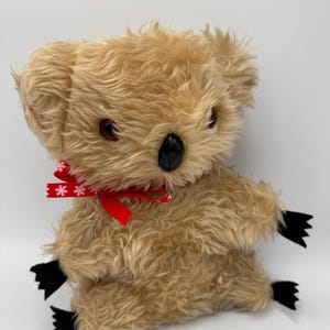 Può includere: Un koala di peluche con pelliccia marrone chiaro, occhi rossi e naso nero. Ha zampe nere e un fiocco rosso con fiocchi di neve bianchi. Il koala è un peluche.