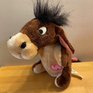 Może przedstawiać: Pluszowa zabawka osioł w kolorze brązowym i beżowym z czarną grzywą i różowymi uszami. Zabawka ma różowy guzik na brzuchu z napisem "Droopee".
