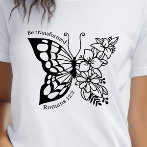 以下が含まれることがあります： 黒い蝶と花のデザインが施された白いTシャツ。蝶には「Be transformed」と「ローマ人への手紙 12:2」の引用が印刷されています。