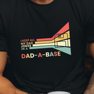 Op de afbeelding: Zwart T-shirt met de tekst "I keep all my dad jokes in a DAD-A-BASE" in een kleurrijk geometrisch ontwerp. Het shirt is gemaakt van zacht materiaal, ideaal voor Vaderdag.