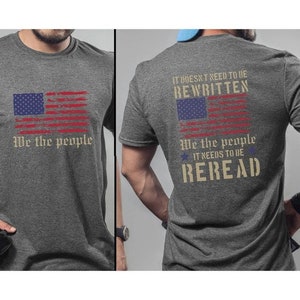 Könnte beinhalten: Ein dunkelgraues T-Shirt mit einem abgenutzten amerikanischen Flaggen-Design und dem Text "We the people" auf der Vorderseite. Die Rückseite des Shirts zeigt den Text "It doesn't need to be rewritten. We the people. It needs to be reread." mit einem abgenutzten amerikanischen Flaggen-Design.