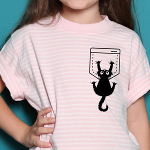 Peut inclure: Un t-shirt rayé rose et blanc avec un chat noir qui sort d'une poche. Le chat regarde vers le haut et a une expression surprise.