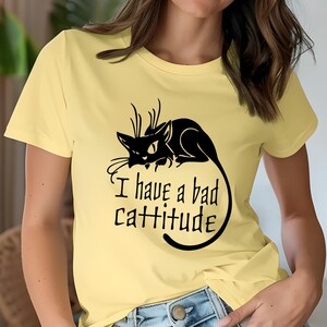 May include: A light yellow t-shirt featuring a black cat graphic with the text "I have a bad cattitude". The cat is lying down with its tail curled. The shirt is a crew neck style.