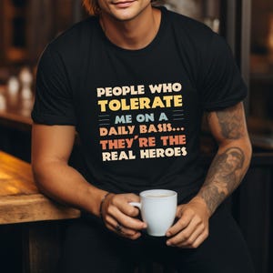 May include: Black crew neck t-shirt featuring the text "PEOPLE WHO TOLERATE ME ON A DAILY BASIS... THEY'RE THE REAL HEROES" in yellow, blue, and red. The shirt is a casual style.