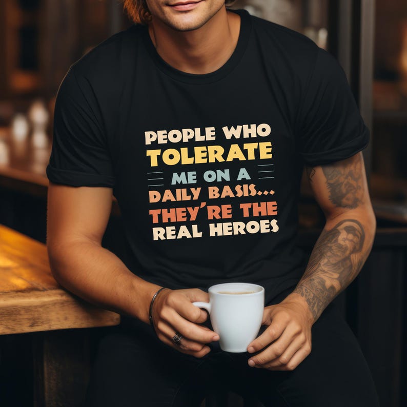 May include: Black crew neck t-shirt featuring the text "PEOPLE WHO TOLERATE ME ON A DAILY BASIS... THEY'RE THE REAL HEROES" in yellow, blue, and red. The shirt is a casual style.
