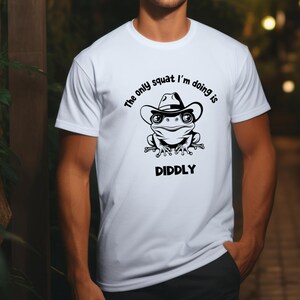 May include: White t-shirt with a black and white illustration of a frog wearing a cowboy hat. The text "The only squat I'm doing is DIDDLY" is printed on the shirt.