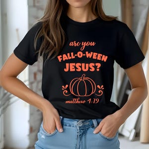 May include: A black short-sleeved t-shirt featuring the orange text "are you FALL-O-WEEN JESUS?" above a graphic of a pumpkin with decorative swirls and the text "matthew 4:19" printed in orange below the pumpkin graphic on the front of the shirt.