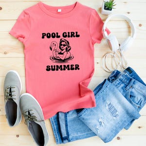 May include: A coral-colored t-shirt with the words "POOL GIRL SUMMER" and a graphic of a woman on a flamingo float. The shirt is paired with blue jeans, gray sneakers, and white headphones.