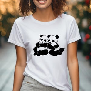May include: White t-shirt featuring a black and white graphic of two hugging pandas. The pandas are depicted in a simple, cartoon style, with black bodies and white faces. The shirt is made of a soft, comfortable material.