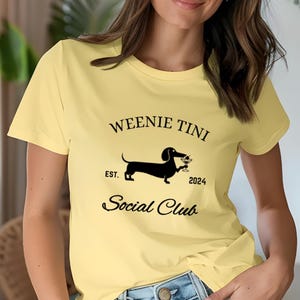 Pode incluir: Camiseta amarela com um dachshund preto segurando uma taça de martini. O texto na camiseta diz "Weenie Tini Social Club Est. 2024".