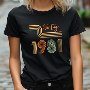 May include: Black t-shirt with a retro design. The shirt features the word "Vintage" in orange script above the number "1981" in a retro font. The design includes orange, green, and brown stripes.