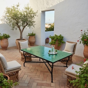 Op de afbeelding: Buitenpatio set met een groene betegelde tafel en rotan stoelen met crèmekleurige kussens. De tafel heeft een zwart metalen frame. Potplanten en een kleine olijfboom dragen bij aan de sfeer. De patio staat op een bakstenen ondergrond.