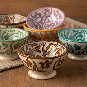 Könnte beinhalten: Eine Sammlung von sechs Keramikschalen mit aufwendigen Blumenmustern in verschiedenen Farben. Die Schalen haben eine cremefarbene Basis mit Designs in Braun, Grün, Lila und Türkis. Jede Schale hat einen weißen Sockel und einen kontrastierenden Rand.