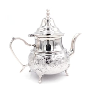 Op de afbeelding: Een zilveren theepot met een versierd ontwerp en drie poten. De theepot heeft een deksel met een knop bovenop.