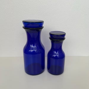 May include: Two cobalt blue glass jars with matching lids. The larger jar features a wide base and a tapered neck, while the smaller jar has a similar cylindrical shape. Both containers are made of smooth, translucent blue glass with flat tops.