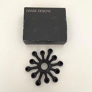 Op de afbeelding: Een zwarte, stervormige kandelaar met meerdere cirkelvormige openingen voor kaarsen. De kandelaar staat naast een zwarte doos met de woorden "DANSK DESIGNS" erop.