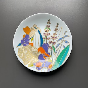 Op de afbeelding: Een wit keramiek bord met een bloemenontwerp. De bloemen zijn geschilderd in tinten paars, oranje, geel en groen. Het bord heeft een gouden rand en een gouden bloemenontwerp.