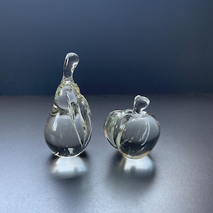 May include: Two clear glass fruit figurines, one shaped like a pear with a tall stem and one shaped like an apple with a small stem. Both pieces are made of smooth, transparent glass that reflects light, creating bright highlights on their surfaces.
