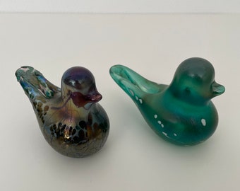 Könnte beinhalten: Zwei Glasvogel-Figuren. Eine ist grün mit weißen Flecken und die andere ist schillernd mit braunen und violetten Akzenten.