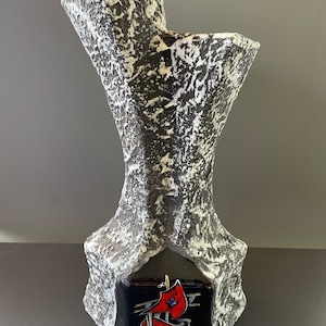 May include: A black and white ceramic vase with a textured surface. The vase has a unique shape with a wide opening at the top and a narrow base. The base of the vase features a black and blue tile with a red and white sailboat design.