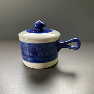 Op de afbeelding: Een witte keramische pot met een blauw deksel en handvat. De pot heeft een blauw en wit bladpatroon rond het midden.