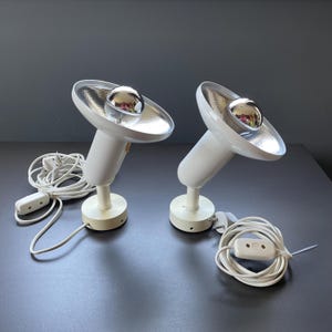 Op de afbeelding: Twee witte bureaulampen in retrostijl, met een zilveren reflector en een chromen lamp. Elke lamp heeft een ronde voet en een snoer met een aan/uit-schakelaar. De lampen staan op een donkere ondergrond.