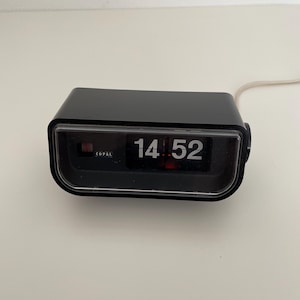 Puede incluir: Un reloj de solapa rectangular negro de marca Copal con una pantalla frontal transparente que muestra la hora 14:52 en números blancos. El reloj presenta un diseño horizontal con un cable de alimentación fino y una perilla de ajuste lateral.