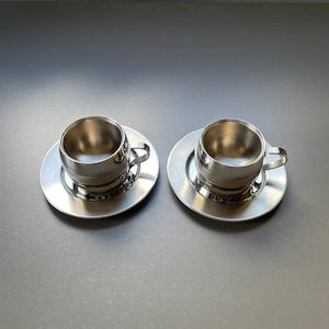 Puede incluir: Dos tazas de espresso de acero inoxidable con sus platillos a juego. Las tazas tienen forma redondeada con asas pequeñas. Los platillos son planos y circulares. El juego está hecho de metal brillante de color plateado.
