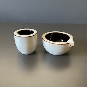 May include: Two white ceramic bowls with brown rims. One bowl is round and the other is a small, cylindrical shape.