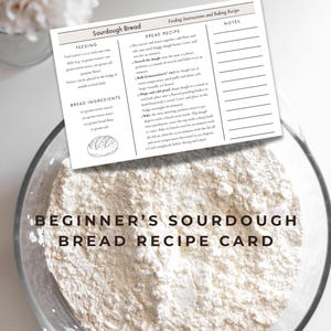 Pode incluir: Uma tigela de vidro transparente cheia de farinha, com um cartão de receita em cima. O cartão diz "Beginner's Sourdough Bread Recipe Card" e inclui instruções e ingredientes. Um pequeno vaso de flores brancas está no fundo.