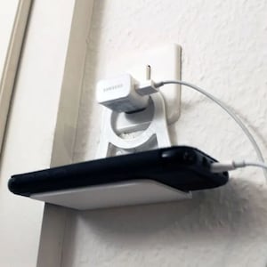 Könnte beinhalten: Weiße Wandhalterung aus Kunststoff mit einem Telefonladeständer und einem Samsung-Ladegerät, das an eine Steckdose angeschlossen ist. Ein schwarzes Telefon liegt auf dem Ständer.