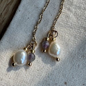 Könnte beinhalten: Hängeohrringe mit Süßwasserperlen und kleinen lila Perlen, die an zarten Goldketten hängen. Die Ohrringe haben goldfarbene Akzente und werden auf einem neutralen Leinenhintergrund präsentiert.