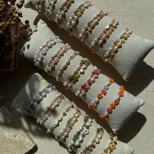 Könnte beinhalten: Eine Sammlung von Perlen- und Blumenarmbändern in verschiedenen Farben, darunter Weiß, Rosa, Orange, Grün und Blau. Die Armbänder werden auf einer weißen Stoffoberfläche präsentiert.