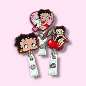 May include: Three glittery badge reels featuring Betty Boop designs. One is heart-shaped with Betty and a dog, one shows Betty's face with gold earrings, and one depicts Betty sitting on red cherries. Each has a white plastic clip and metal hardware.