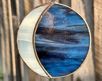 May include: Stained glass crescent moon suncatcher with a celestial design. The moon features a white, textured crescent shape and a blue, swirling night sky design. It hangs from a silver chain and ring.