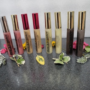 May include: Eight lip gloss tubes with gold caps and clear plastic tubes. The tubes are labeled with the brand name "DEWE NATURALS" and the flavor or color of the gloss. The flavors are Pink Candy, Champagne Teaser, Naturally Clear, and Balmy Brown.