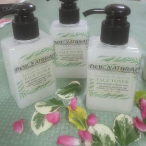 May include: Three clear glass bottles of face toner with white labels. The labels have the text "DEW NATURALS" and "CLARIFYING FACE TONER" with different scents: Lavender & Rose, Lavender & Jasmine, and Lavender & Jasmine. The bottles are on a green and white surface.