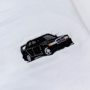 Puede incluir: Tela blanca con un pequeño diseño de coche negro bordado. El coche está detallado con ruedas plateadas y faros amarillos. La tela es lisa, posiblemente de algodón o un material similar.