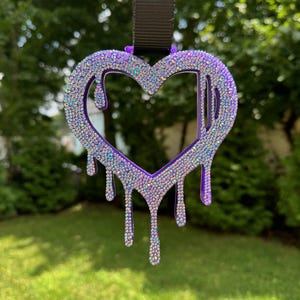 Puede incluir: Un amuleto de corazón morado con un diseño de goteo, cubierto de diamantes de imitación iridiscentes. El amuleto está unido a un cordón negro.