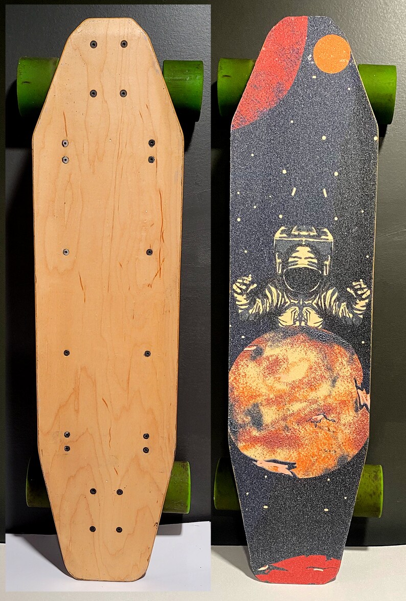 May include: A skateboard deck with a wood grain finish on one side and a space-themed graphic on the other. The graphic features an astronaut standing on a planet with a red and orange color scheme. The background is black with white stars.