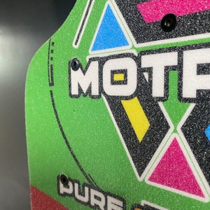 May include: Green skateboard deck with black, white, pink, blue, and yellow geometric shapes and the word "MOTIF" in white. The deck also has the words "PURE SYN" and "VIT" in white.