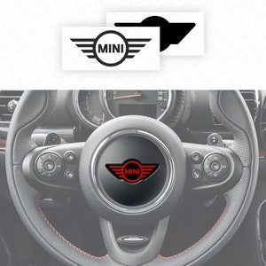 Può includere: Un volante nero con un logo MINI rosso al centro. Il volante ha dettagli argentati e pulsanti sui lati sinistro e destro.